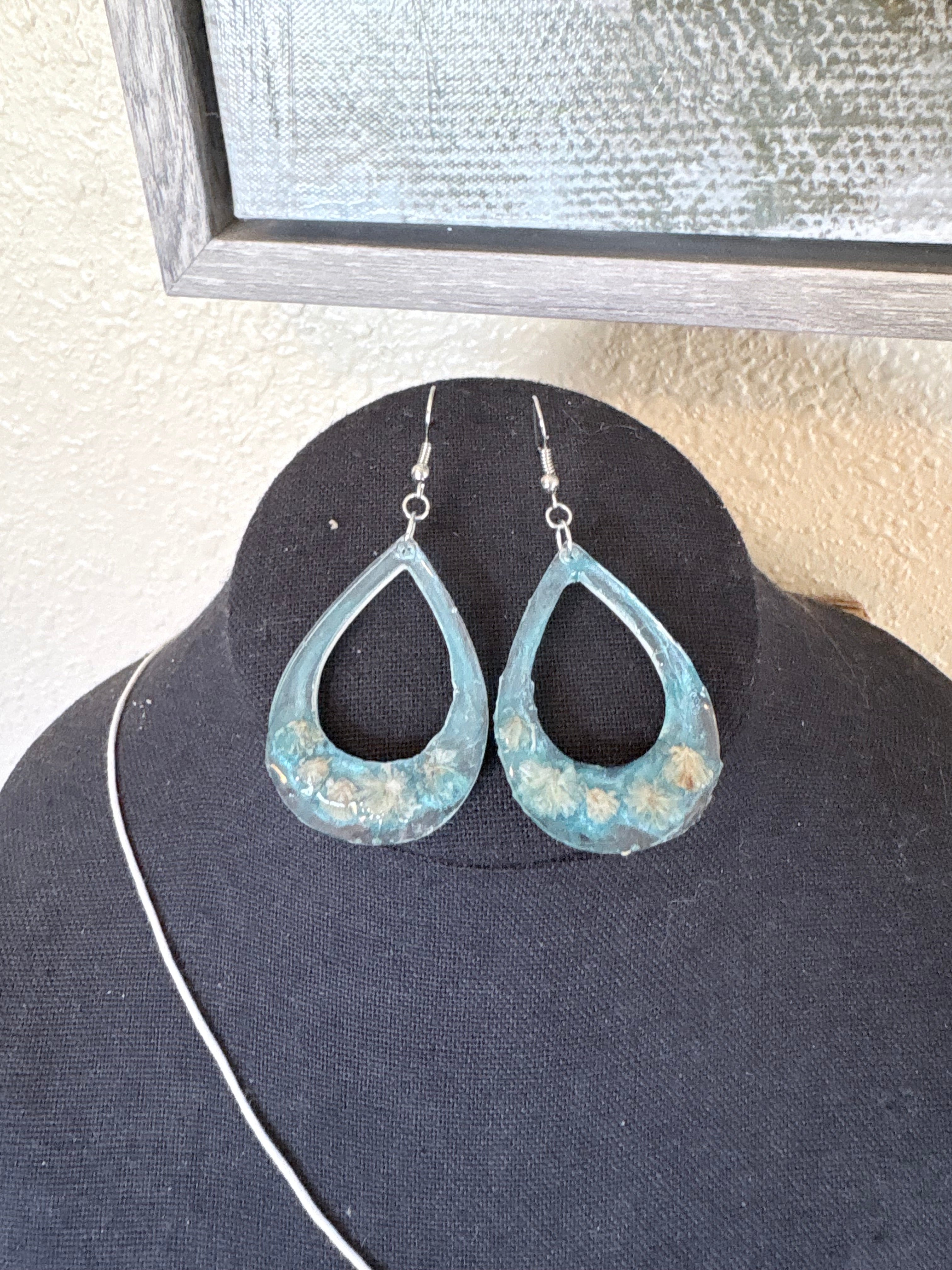 A pair of teardrop-shaped dangling earrings with a distressed blue resin and white flowers, displayed on a black jewelry bust.