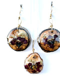 A pair of dangling earrings and a matching pendant featuring round natural wood slices with embedded dried flowers in burgundy and gold, suspended from gold hooks against a white background.
