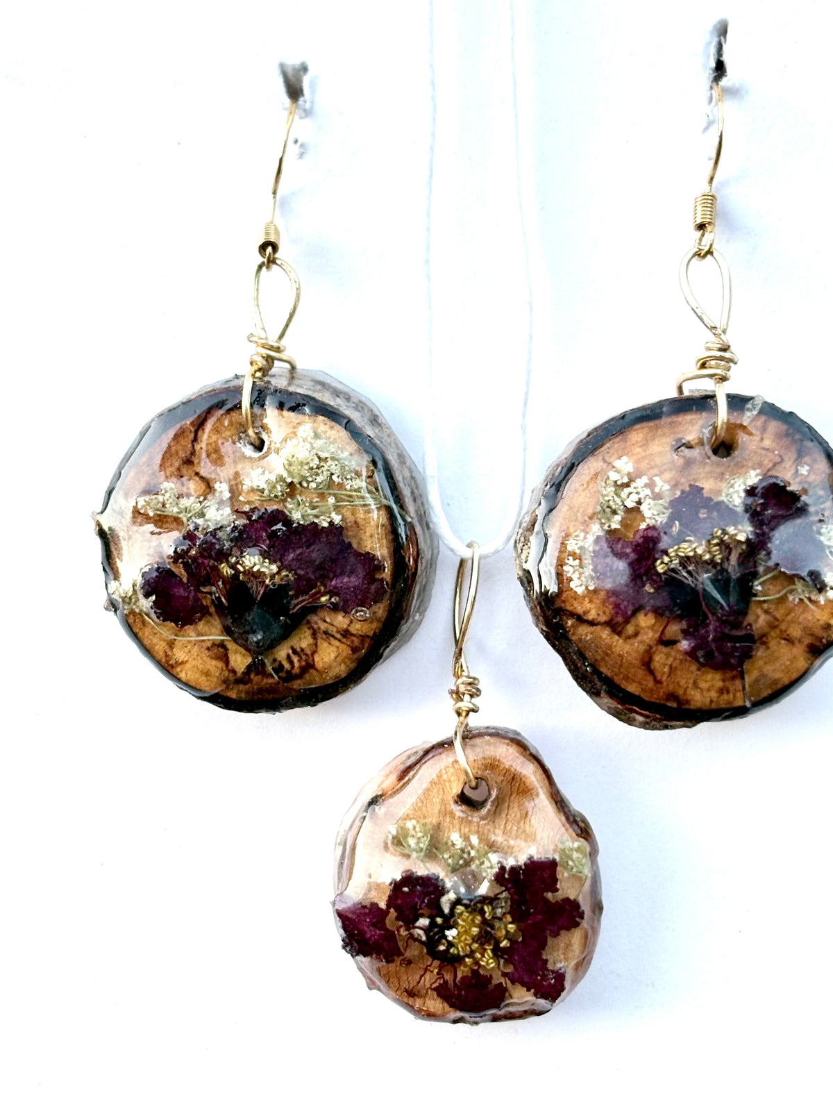 A pair of dangling earrings and a matching pendant featuring round natural wood slices with embedded dried flowers in burgundy and gold, suspended from gold hooks against a white background.