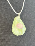A teardrop-shaped pendant necklace made of translucent light green resin with a small pink flower encased inside, hanging from a white cord against a dark textured fabric background.