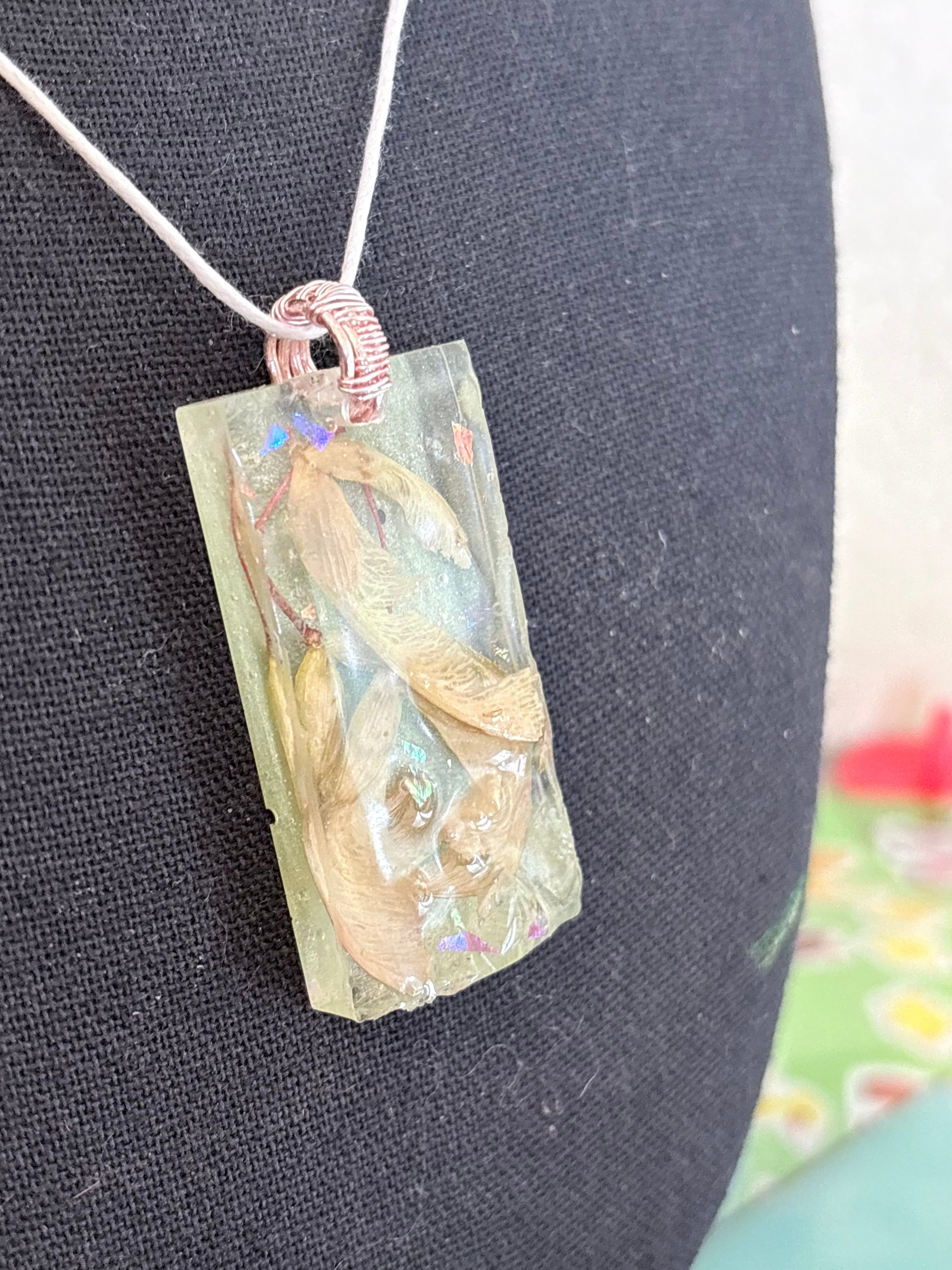 A handmade rectangular resin pendant necklace containing dried plant seeds, suspended from a white cord with a copper-colored wrapped bail, displayed on a black fabric jewelry bust.