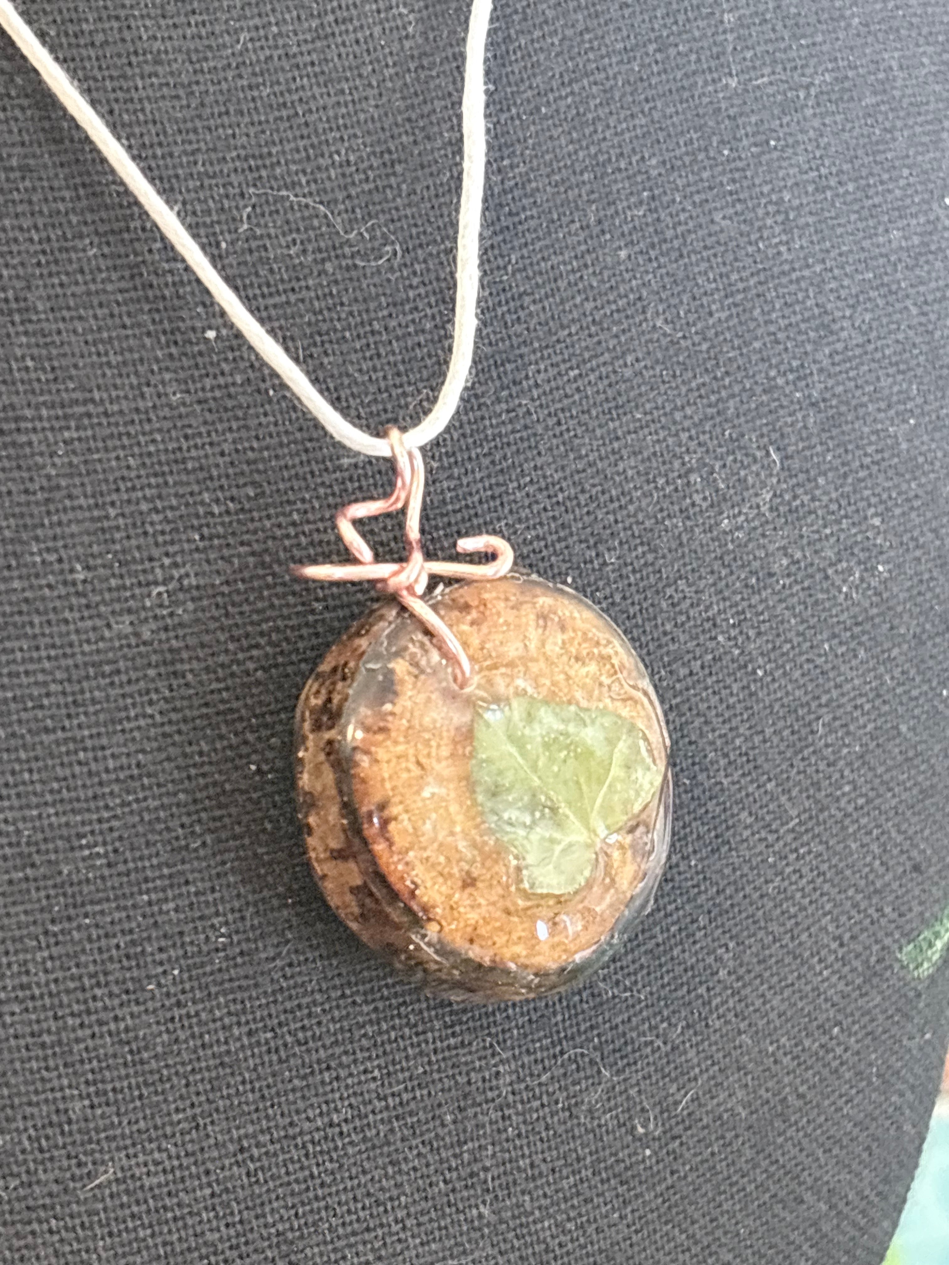 A handmade pendant necklace featuring a round resin wood slice containing a small green leaf, wrapped in copper wire at the top with a white cord, displayed on a black fabric background.