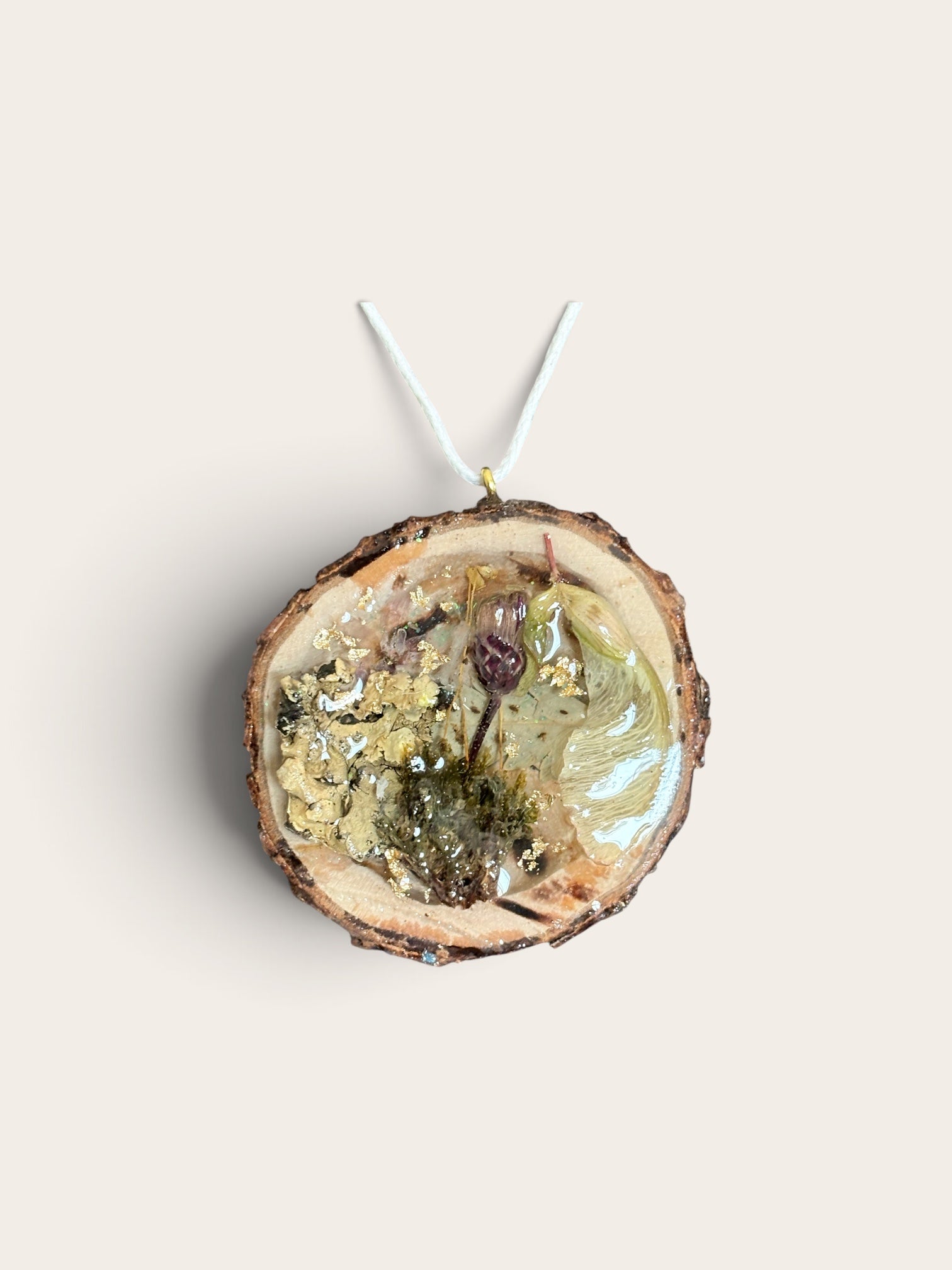 A circular resin pendant made from sliced wood, featuring dried botanical elements inside, suspended by a white string against a solid light beige background.