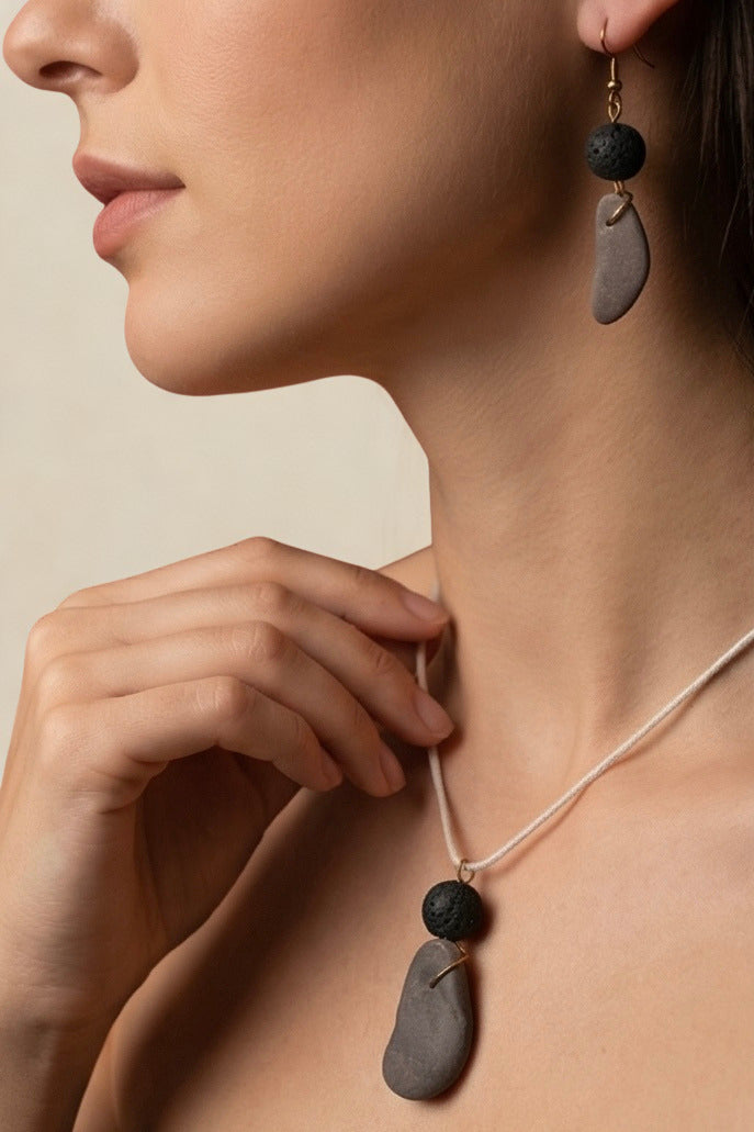 Close-up of a model wearing a minimalist necklace and matching drop earring. The jewelry features smooth, irregular gray stone pendants and dark lava beads on a thin cream cord, set against a warm beige background.