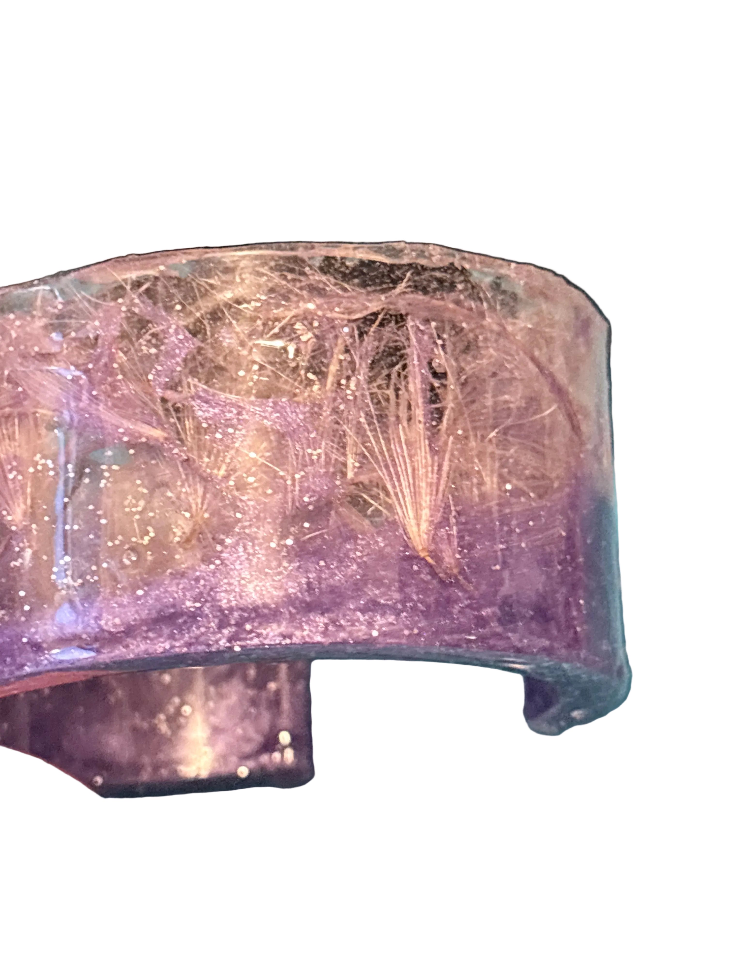 A close-up of a handmade resin cuff bracelet containing visible thistle strands and glitter, held over white background.