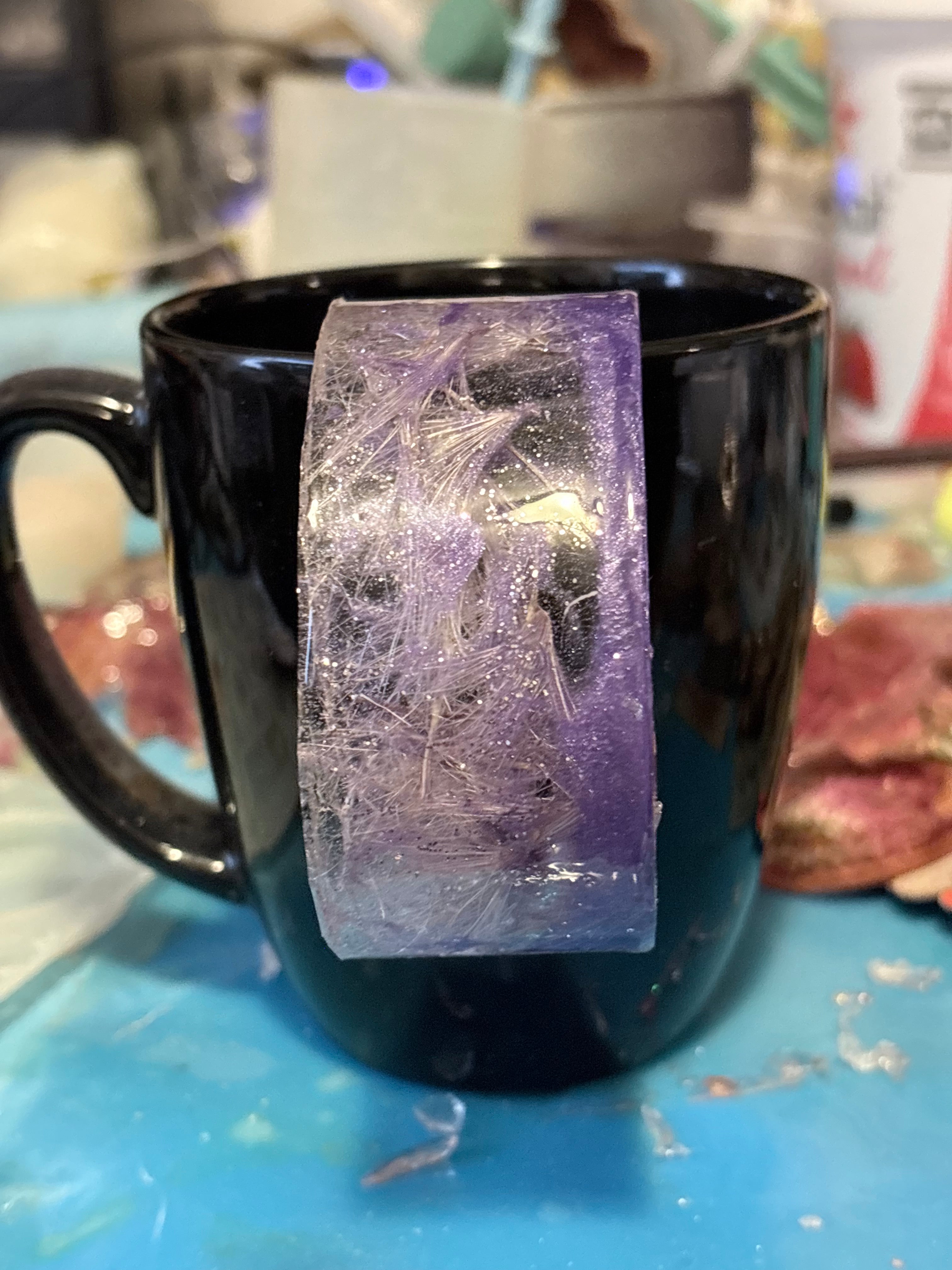 A black ceramic mug with a rectangular translucent purple and clear and purple resin bracelet hung on the outside, set on a blue surface with a blurred workshop background.