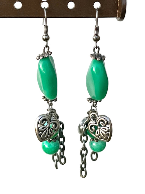 Pair of earrings with green beads and silver heart designs hanging on a hook.